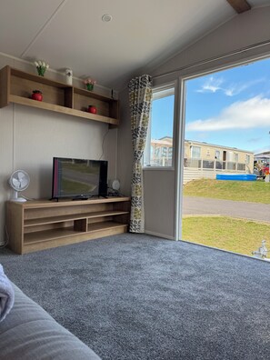 A modern living room with a flat-screen TV on a wooden stand, a white fan, and a grey carpet. A large window offers a view of a caravan park. - Close to the beach (Leysdown on sea)