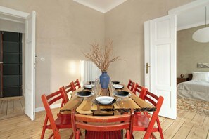 Comedor con mesa de madera dispuesta para seis personas, rodeada de sillas rojas, un florero azul con ramas y una puerta blanca al fondo.