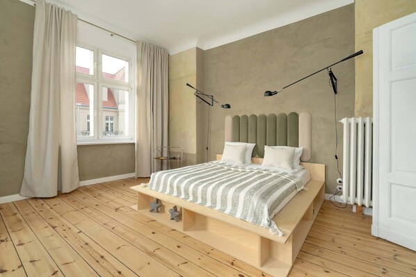 Un dormitorio con piso de madera, una cama con manta a rayas, cabecera, ventana con cortinas y un radiador.