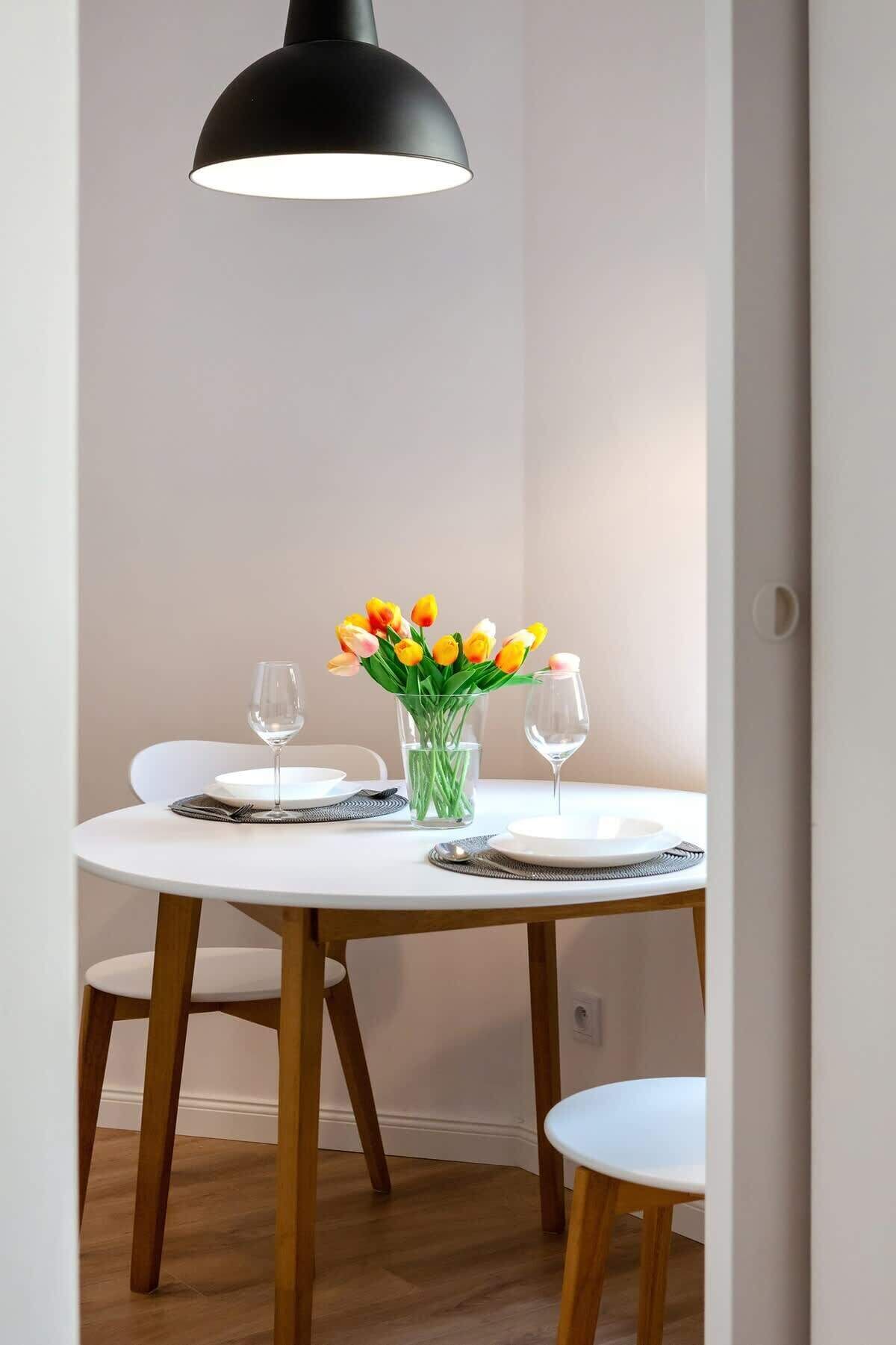 A dining area with a round table, two chairs, a vase of tulips, two wine glasses, and a black pendant light.