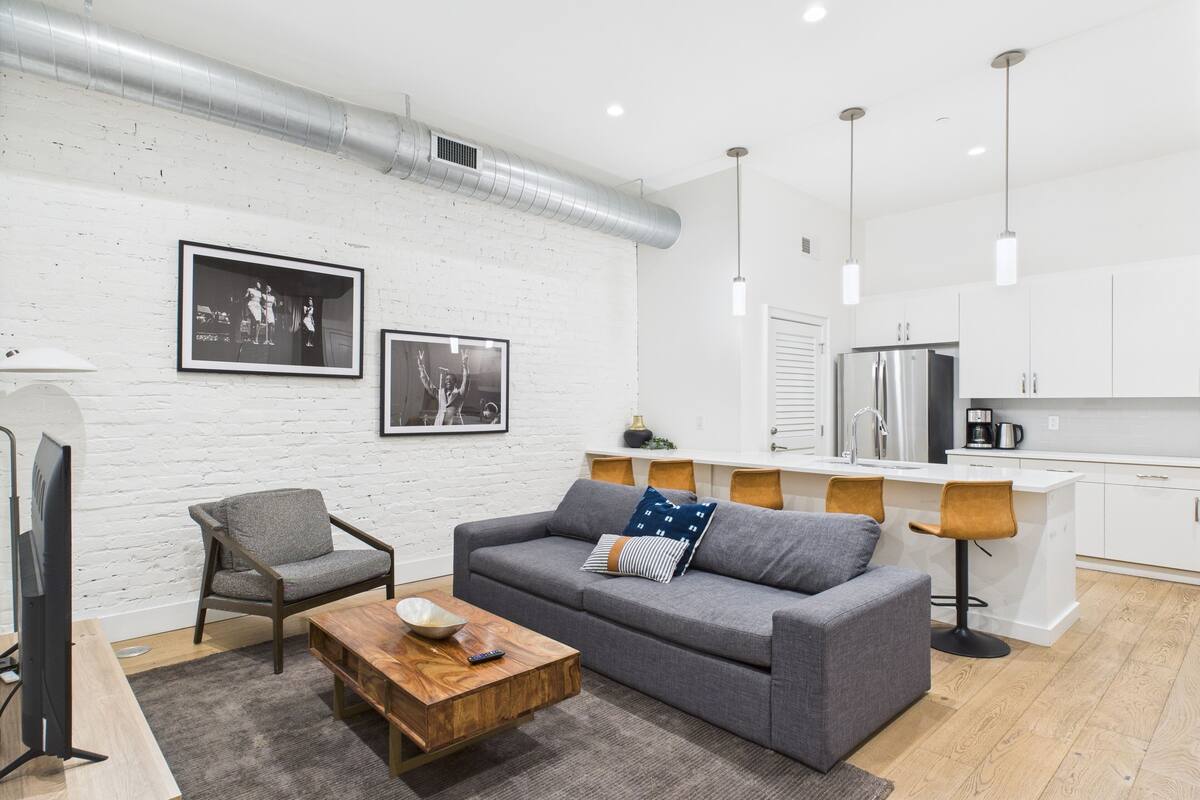Ein modernes Wohnzimmer mit einem grauen Sofa, einem Holztisch und einem Fernseher. Es gibt eine Küchenzeile mit weißen Schränken und einem Edelstahl-Kühlschrank.