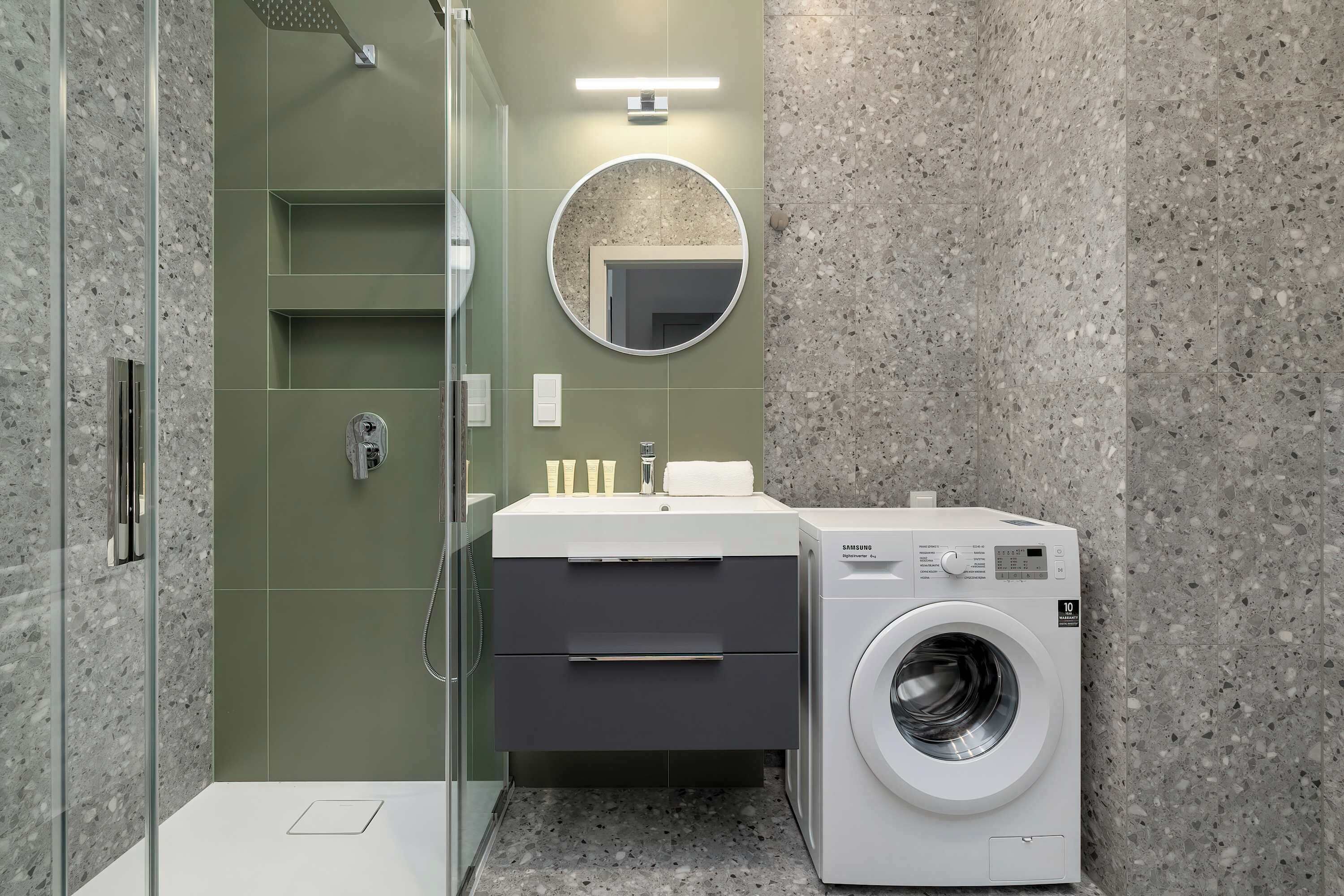Un baño moderno con ducha envuelta en vidrio, un lavamanos con espejo y una lavadora de carga frontal.