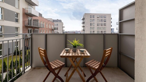 A balcony with a wooden table and chairs, a potted plant, and a view of surrounding buildings. - Bright Apartment with Balcony | Parking, Workspace (Katowice)