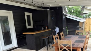 Eine überdachte Terrasse mit einem Holztisch und Stühlen, einer kleinen Bar und einem Ventilator.