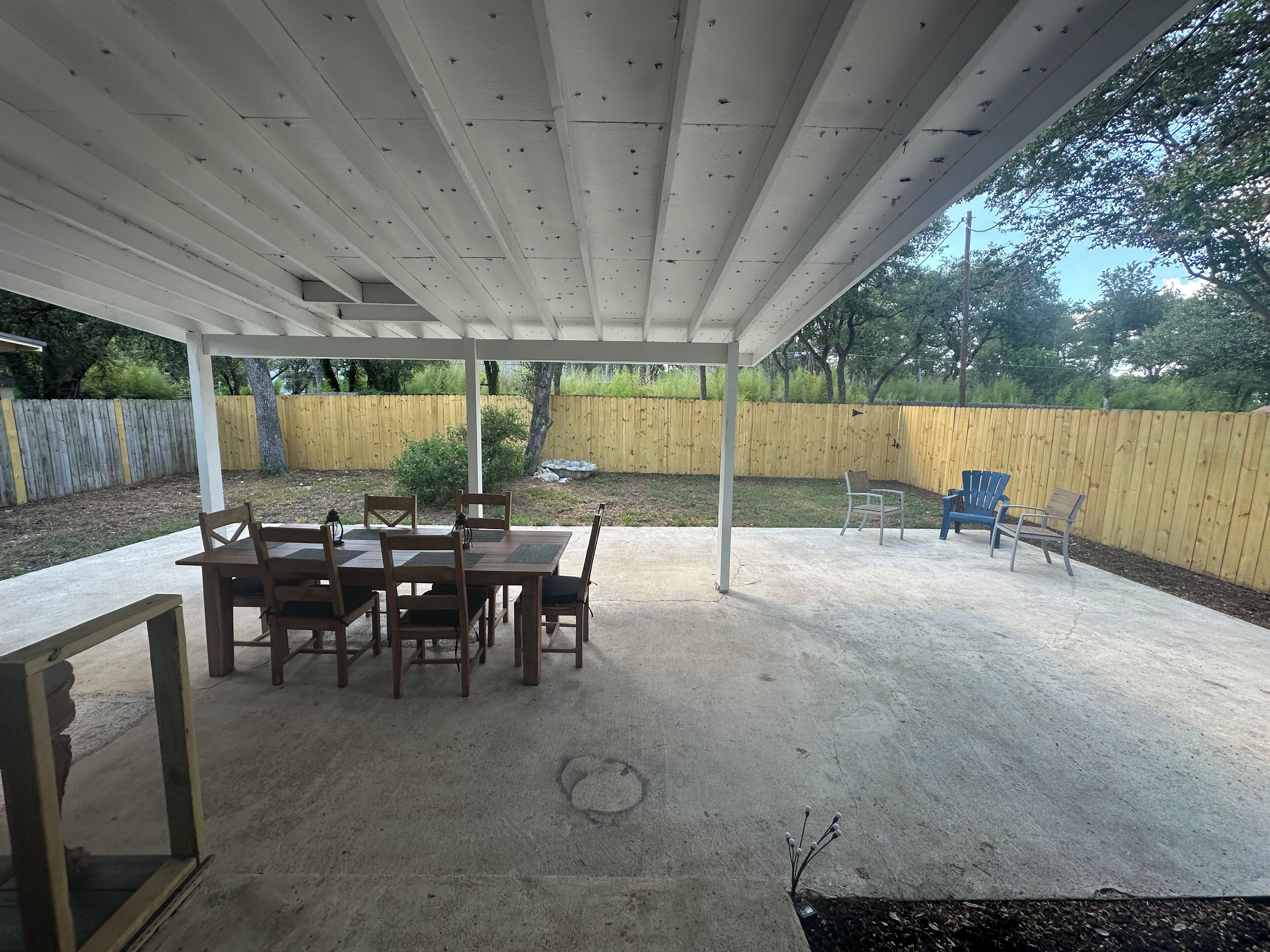 Un patio cubierto con piso de concreto, mesas y sillas de madera, y una cerca amarilla al fondo.