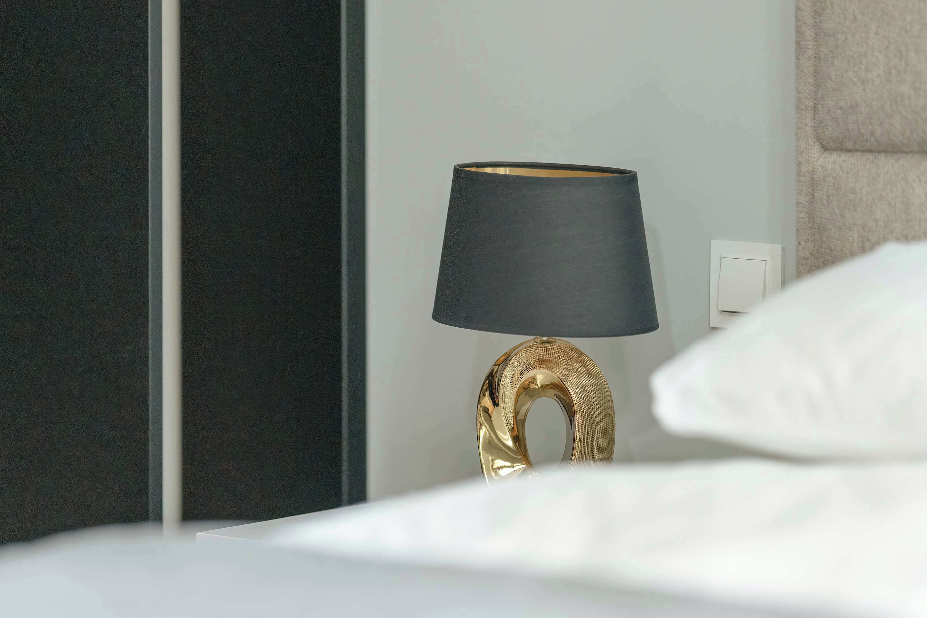 A bed with white linens, a gold-colored lamp with a black shade, and a dark-colored wall.