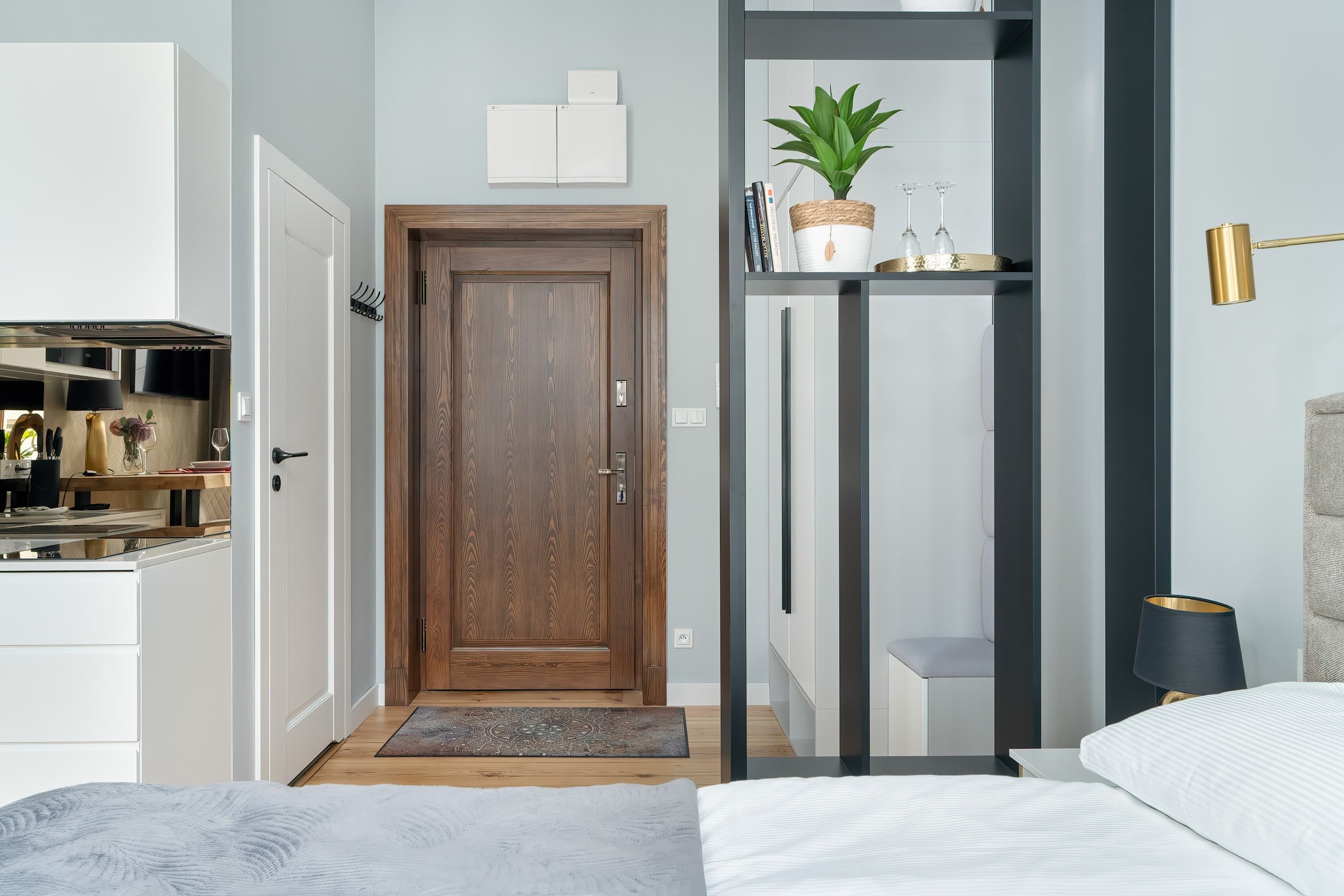Una habitación de hotel moderna con una puerta de madera, un pequeño rincón de descanso con una planta y una mesita de noche con lámpara.