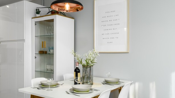 Un comedor moderno con una mesa blanca preparada para cuatro personas, un aparador de vidrio con vajilla y una cita enmarcada en la pared.