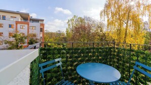 Un balcón con mesa y sillas, rodeado por una cerca verde, con vistas a un edificio y árboles otoñales.