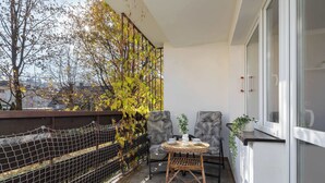 Un balcón con vistas a árboles y edificios, una mesa de mimbre con tazas, dos sillas estampadas y una ventana con persianas.