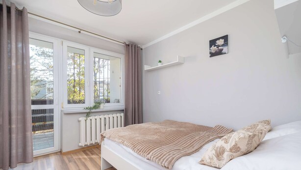 Un dormitorio con cama, ventana con cortinas, radiador y estante en la pared con una planta.