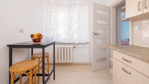 Una cocina pequeña con una mesa, taburetes, un radiador y una ventana con cortinas traslúcidas.
