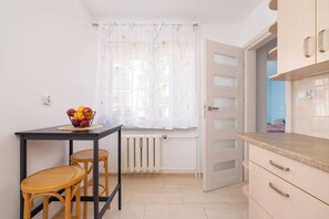 Une petite cuisine avec une table, des tabourets, un radiateur et une fenêtre ornée de rideaux légers.