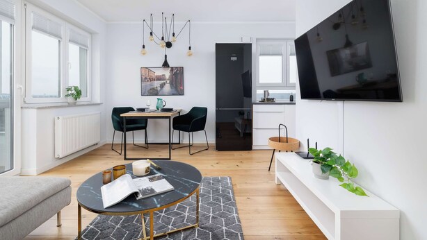Una sala moderna con un sofá, una mesa redonda de centro y un comedor con una mesa y sillas. Un televisor está montado en la pared, y hay una planta en un estante.