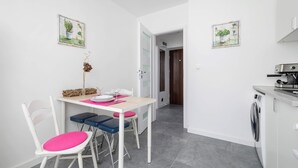 Cozinha compacta com máquina de lavar, mesa de jantar com cadeiras e dois quadros de gravuras botânicas nas paredes.