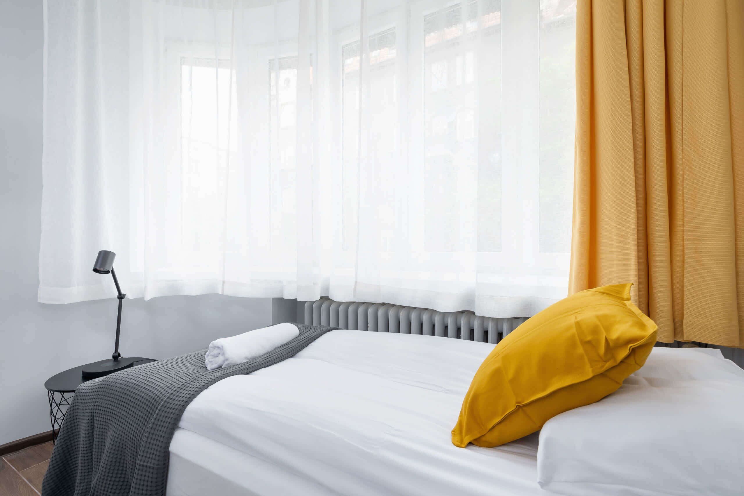 A neatly made bed with a white and gray bedspread, a yellow pillow, and a gray throw blanket. There is a black nightstand with a lamp beside the bed.