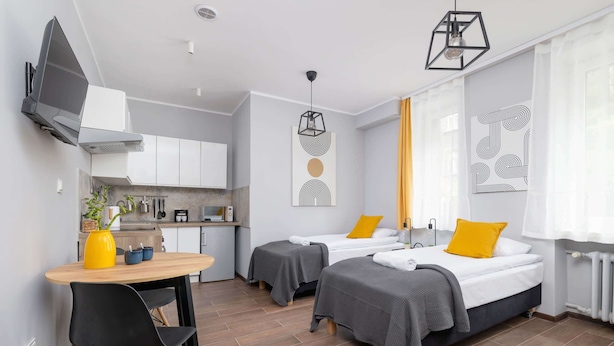 Une chambre d’hôtel moderne dotée d’une kitchenette, de deux lits avec une literie grise, d’une table à manger avec des chaises et d’œuvres d’art accrochées aux murs.