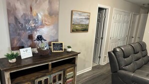 A living room with a dark leather sofa, a wooden console table with a sign, a painting, and a door in the background. - Kayak Cove Lake Lagoon (Grant)