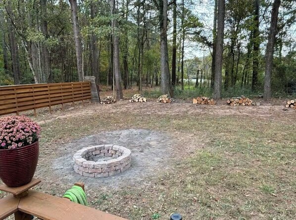 A wooden bench, a large potted plant, a fire pit, and stacks of firewood in a wooded area.