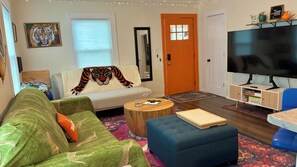 A living room with a sofa, a coffee table, a blue ottoman, a television, and a bookshelf.