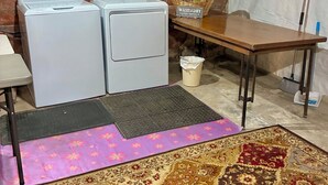 A laundry area with two washing machines, a wicker basket, a table, a broom, and a rug.