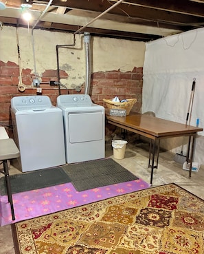 A laundry area with two washing machines, a wicker basket, a table, a broom, and a rug.