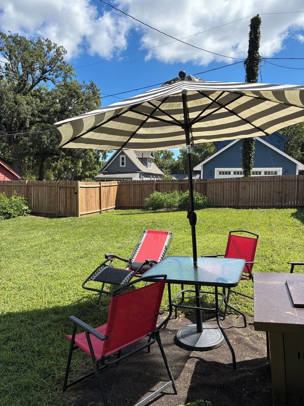 Un patio al aire libre con una mesa, sillas, un gran paraguas y una cerca de madera al fondo.