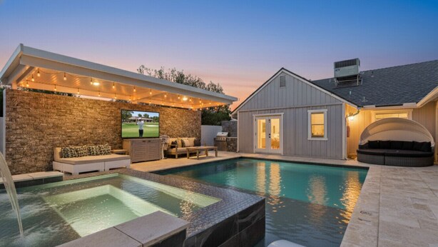 A backyard with a swimming pool, a stone wall, a covered seating area, and a small building with a TV screen. - Scottsdale Oasis | Heated Pool | Spa | Grill (Scottsdale)