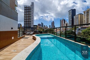 A rooftop swimming pool with a wooden deck and a glass railing, surrounded by high-rise buildings and a clear sky. - Comfortable Studio in Caminho das Árvores (Salvador)