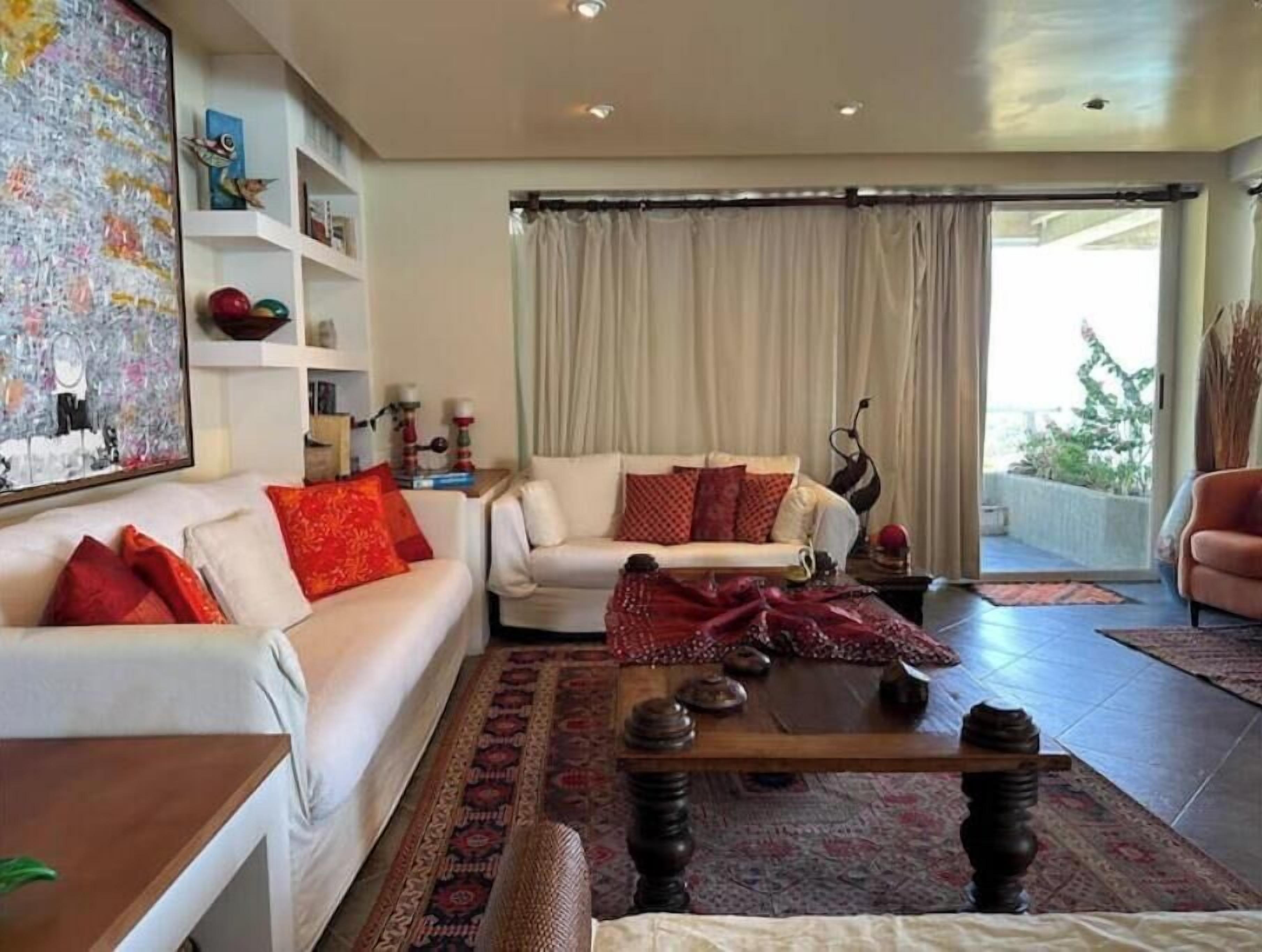Ein Wohnzimmer mit einem weißen Sofa, roten Kissen, einem Couchtisch und einem gemusterten Teppich. An der Wand hängt ein Bild, und ein Bücherregal beherbergt dekorative Gegenstände.