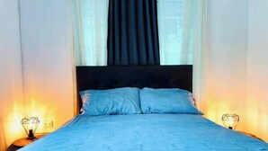 Un lit recouvert d’une couvre-lit bleue, deux lampes de chevet et une fenêtre avec des rideaux.