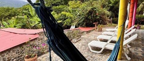 Una hamaca colgada entre dos postes, con un patio empedrado y sillones blancos al fondo.
