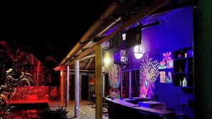 Uma área de bar ao ar livre com estrutura de madeira rústica, letreiro neon e diversas obras de arte nas paredes.