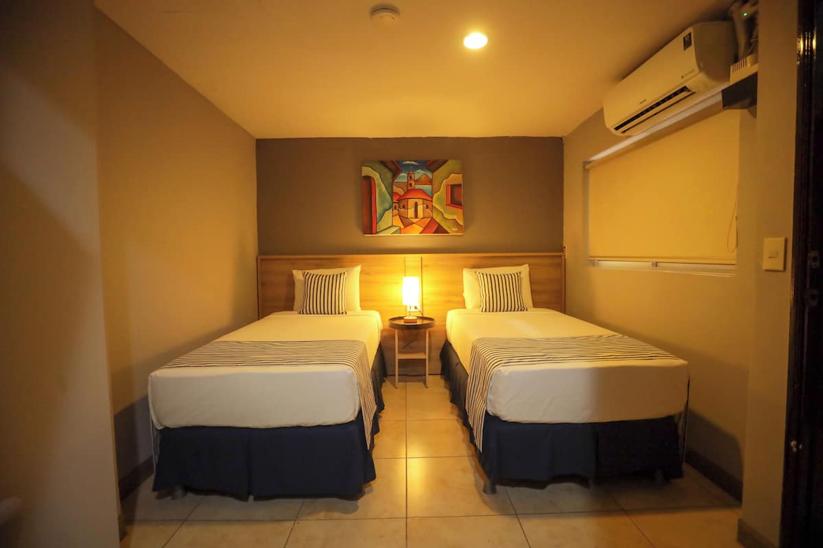 Habitación de hotel con dos camas, una mesita de noche con lámpara y aire acondicionado instalado en la pared.