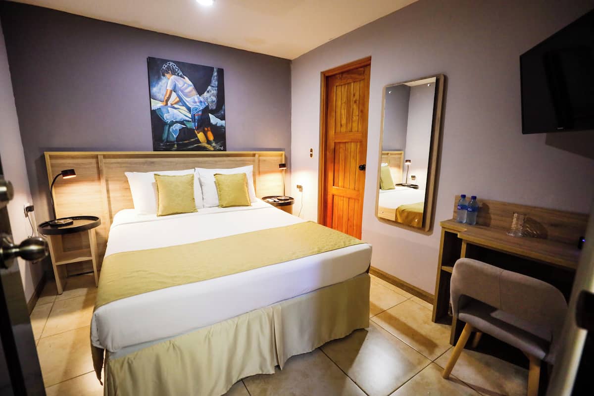 Habitación de hotel con cama, mesitas de noche, escritorio, silla, espejo y un cuadro en la pared.
