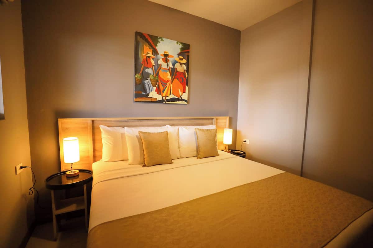 Una habitación de hotel con una cama grande, dos mesitas de noche con lámparas, un cuadro en la pared y una pared lisa.