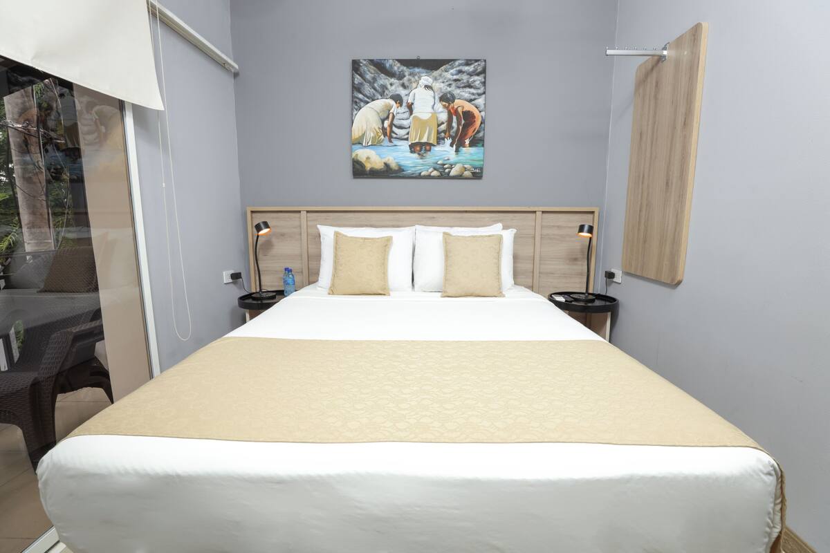Habitación de hotel con una cama grande, dos mesitas de noche, un cuadro colgado en la pared y una ventana con vistas a zonas verdes.