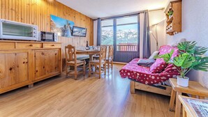 Una habitación acogedora con paredes de madera, una mesa de comedor, sillas, una cama con colcha floral roja y un ventanal con vistas a la montaña.