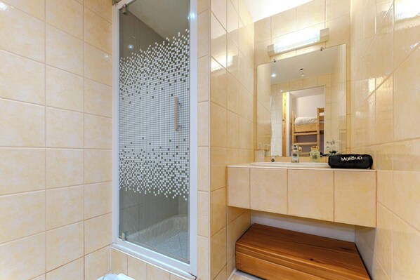 A modern bathroom with a glass shower enclosure, a wooden bench, and a vanity with a mirror and makeup bag.