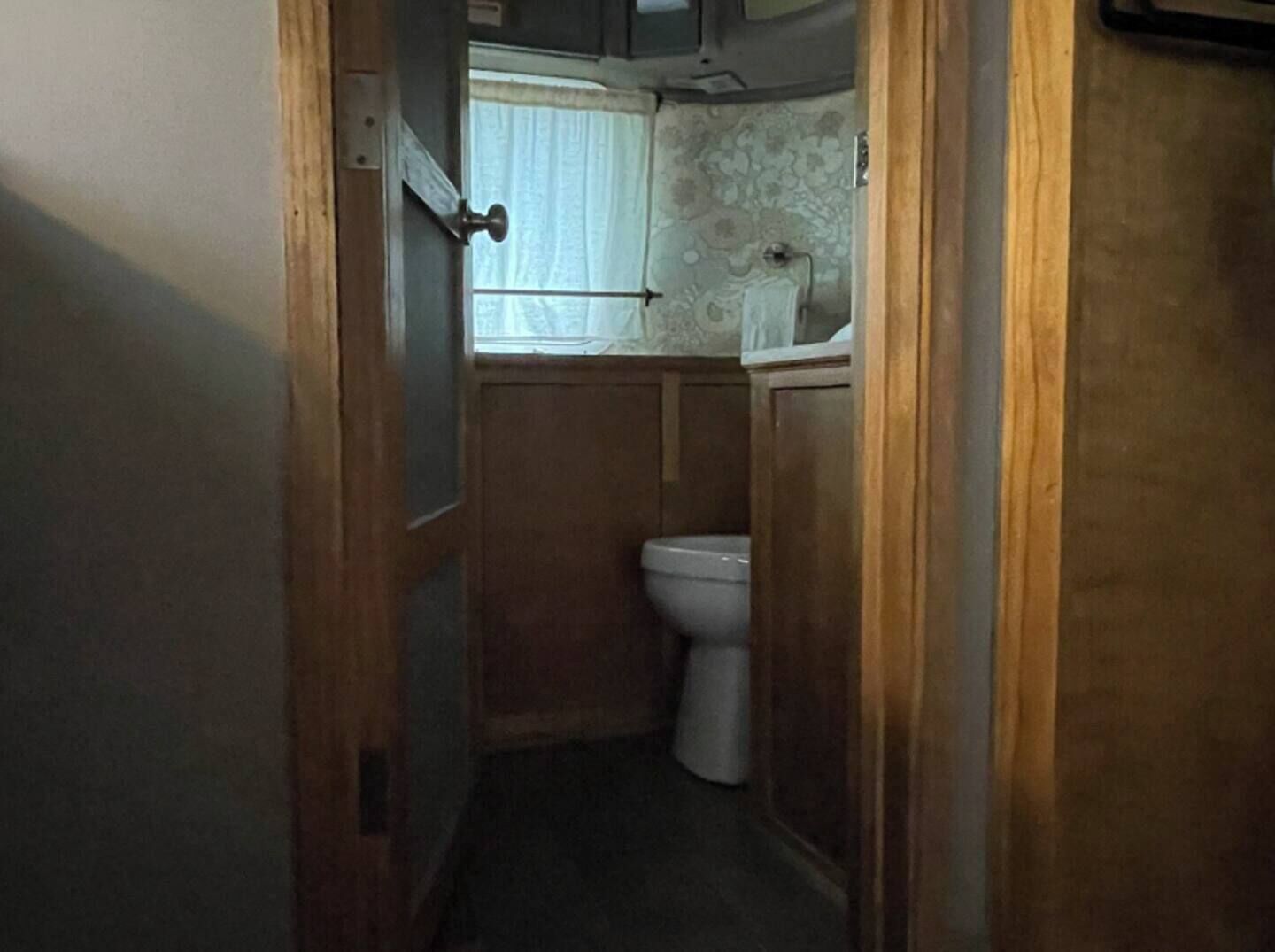 Een smalle gang die leidt naar een kleine badkamer met toilet en een raam met gordijnen.