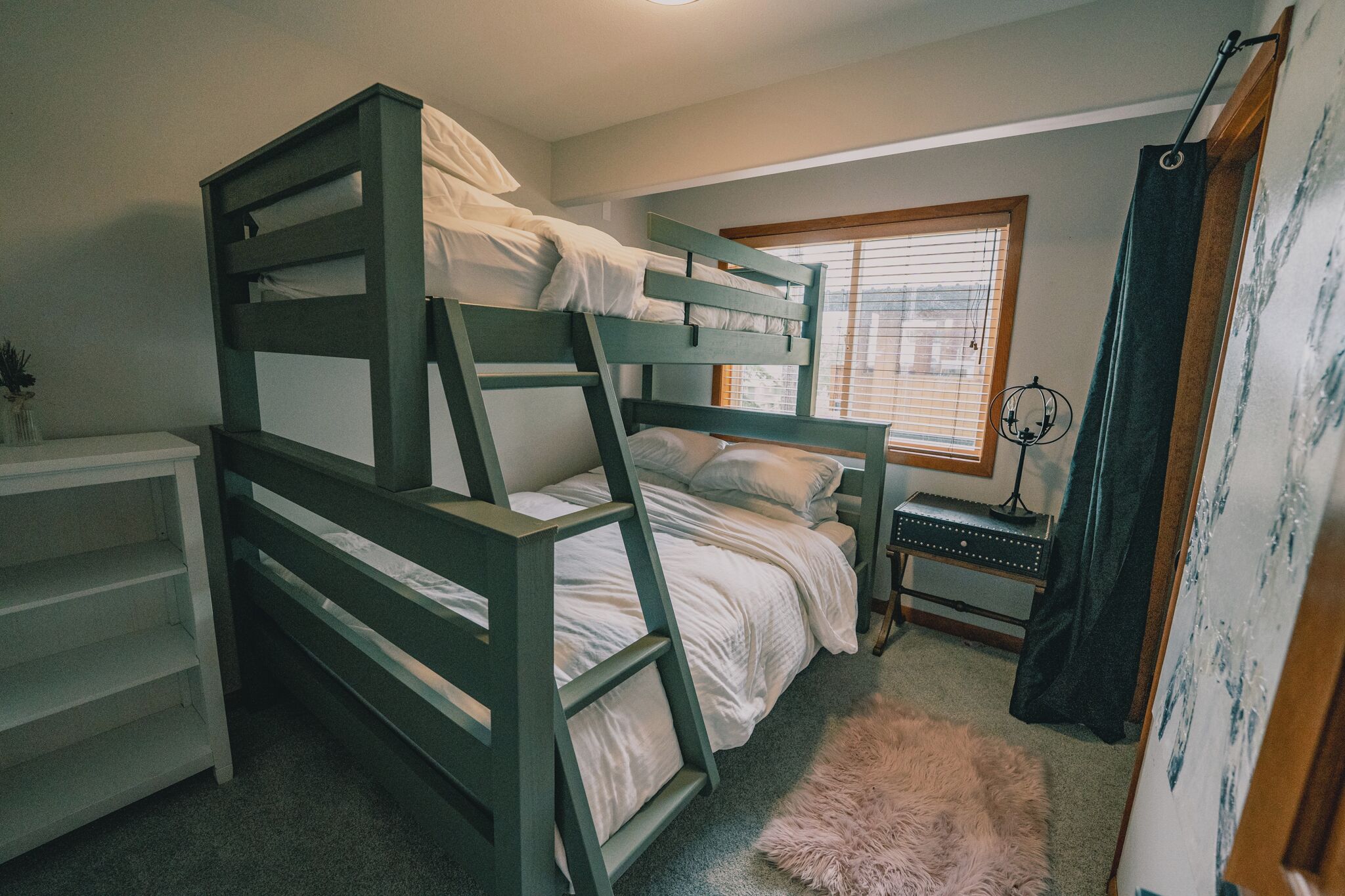Une chambre avec un lit superposé, un bureau, une petite table et une fenêtre avec des stores.