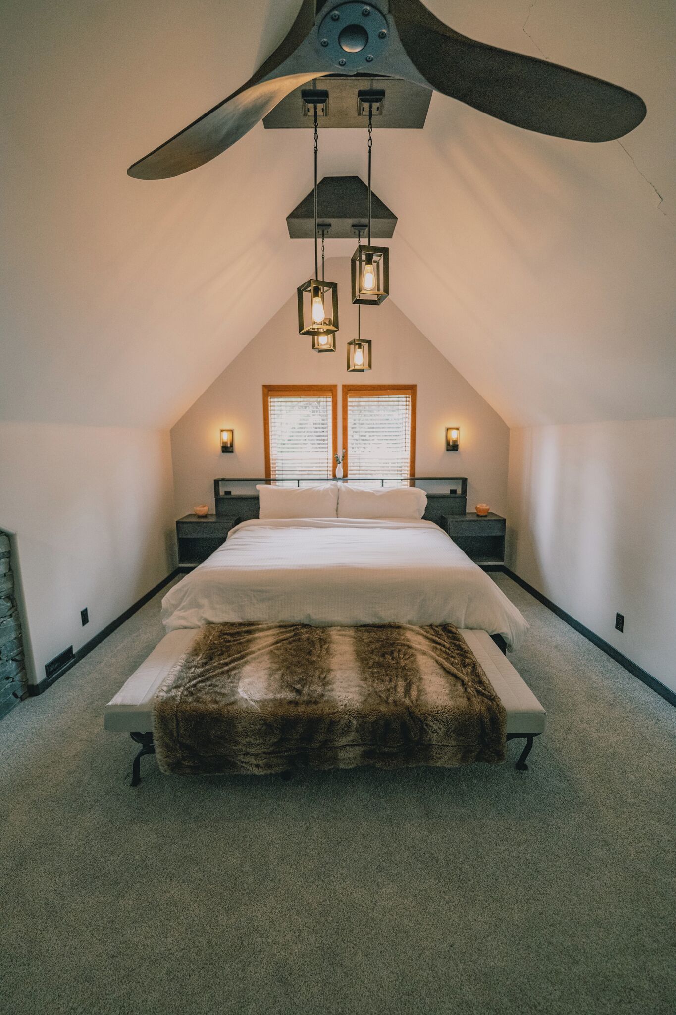 A bedroom with a large ceiling fan, a bed with a fur throw, two bedside tables, and a window with blinds.