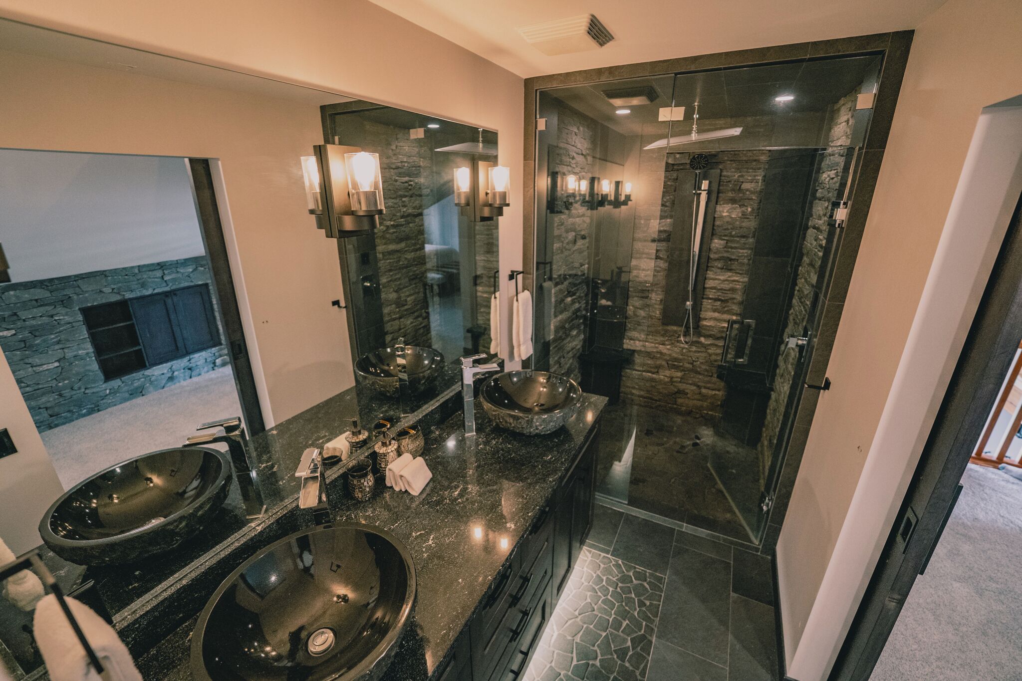 Un baño moderno con dos lavamanos grandes, cabina de ducha de vidrio y pared decorativa de piedra.