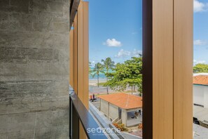 Vista desde un balcón con un muro de concreto y columnas de madera, sobre una playa, palmeras y un pequeño edificio con techo de tejas rojas.