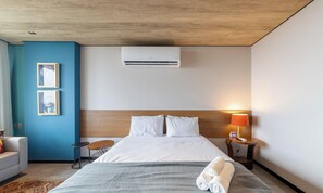 Habitación de hotel con una cama, una mesita de noche con lámpara, una pequeña mesa redonda, una silla y un aire acondicionado de pared.