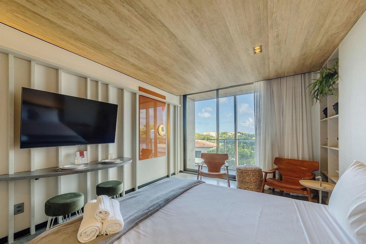 A modern hotel room with a large bed, a flat-screen TV, a small seating area, and a large window offering a city view.
