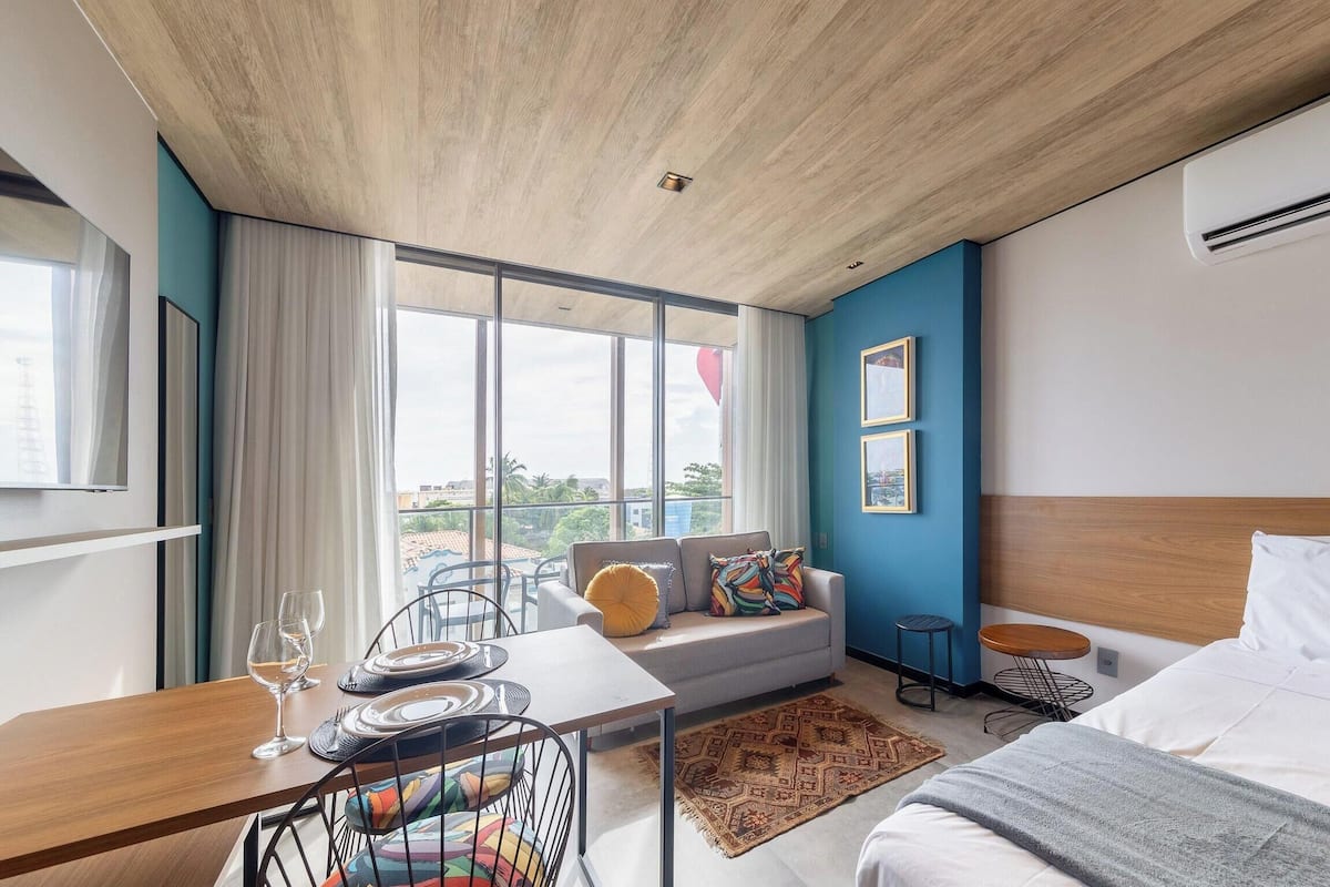 A modern hotel room with a large window, a sofa with colorful cushions, a dining table with plates and a wine glass, a bed with a white duvet, and a small round table.