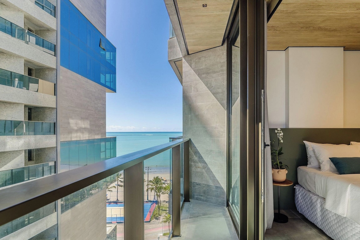 A modern hotel room with a balcony overlooking a beach and a high-rise building.