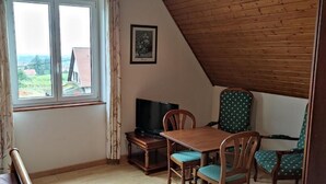 Una habitación con techo de madera, ventana con cortinas, una mesa con sillas, un televisor y un cuadro en la pared.