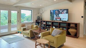 A spacious living room with a high ceiling, a television mounted on the wall, and a large window offering a view of greenery outside. - Centrally Located Bright Family Home with Peaceful Natural Views and Central A/C (Burlington)